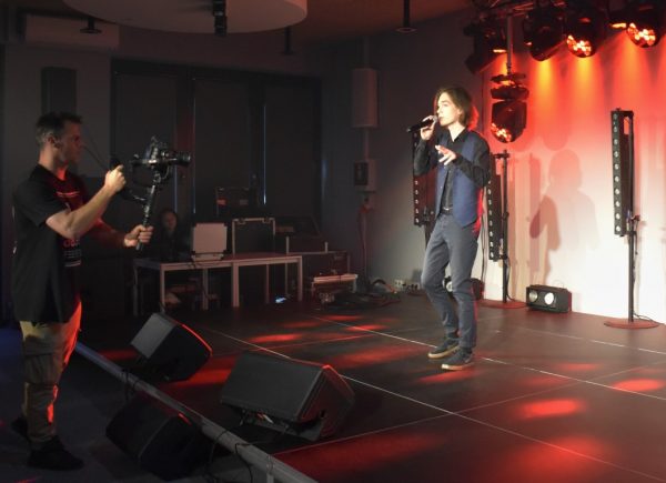 Mężczyzna stoi na scenie. Trzyma w ręku mikrofon i śpiewa. W tle czerwone światło.