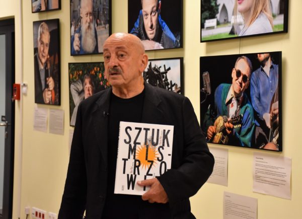 Maciej Kłoś stoi przed ścianą ze zdjęciami znanych osobowości. W rękach trzyma książkę "Sztukmistrzowie".