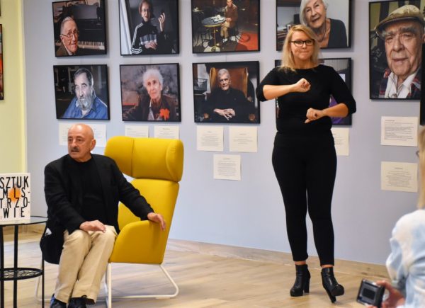 Po lewej stronie na fotelu siedzi Maciej Kłoś. Po prawej stronie kobieta tłumaczy rozmowę na język migowy. W tle na ścianach wiszą zdjęcia z książki "Sztukmistrzowie".