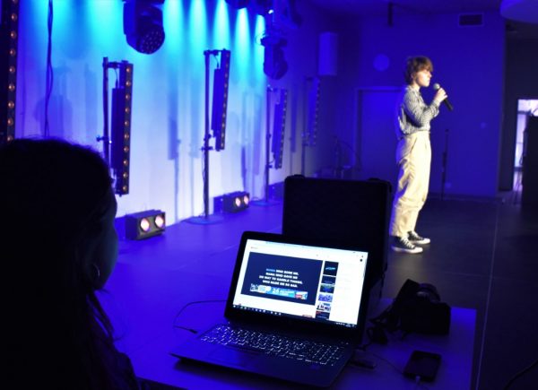 Kobieta stoi na scenie. Trzyma w ręku mikrofon i śpiewa. W tle niebieskie światło. Zdjęcie zrobione zza sceny, widać laptopa i dźwiękowca.