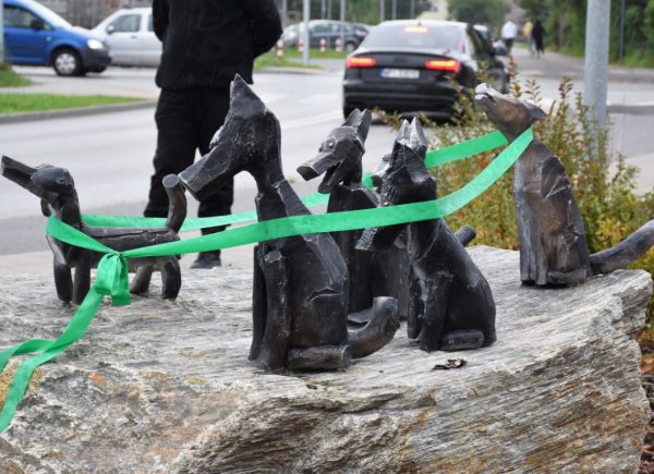 Na zdjęciu widać sześć rzeźb przedstawiających psy. Rzeźby znajdują się na kamieniu. Są one owinięte w zieloną wstążkę zawiązaną na kokardkę.