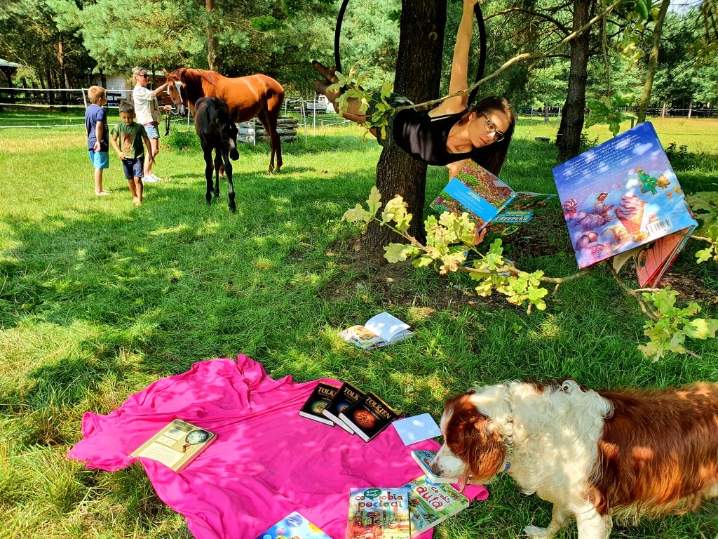 Na zdjęciu widać bajkową scenerię. Kobieta wisi na obręczy i czyta książkę. Na fotografii znajdują się: konie, pies i dziecko. Dziecko czyta książkę na kocu. Jedna publikacja jest zawieszona na drzewie.