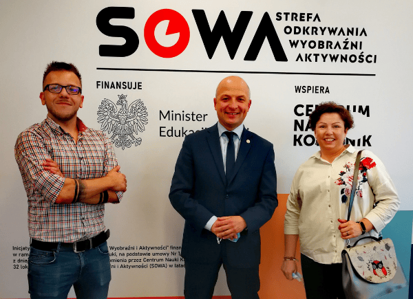 Dyrektor biblioteki Łukasz Załęski z Sylwią Chojnacką-Tuzimek i Danielem Putkiewiczem stoją i pozują do zdjęcia. Za nimi ścianka z banerem promującym inicjatywę SOWA.