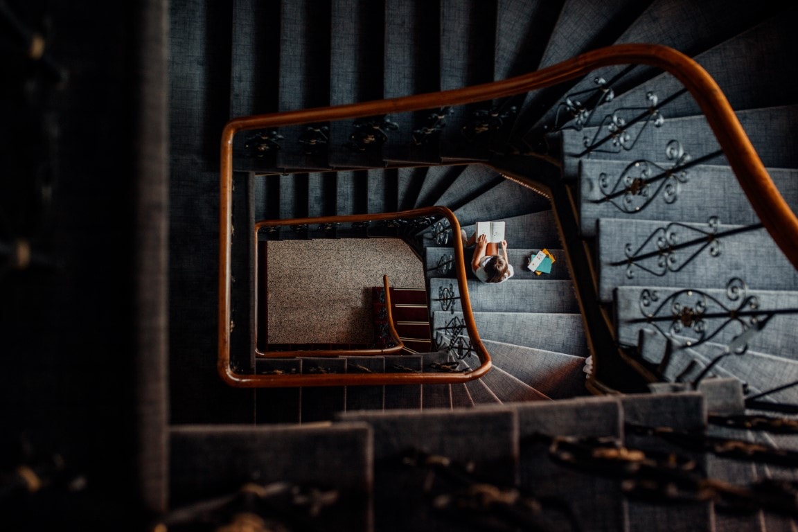 Zdjęcie przedstawia czarne schody. Na końcu schodów siedzi dziewczynka i czyta książkę.