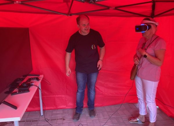 W czerwonym namiocie stoi pracownik biblioteki i pokazuje kobiecie, jak używać okularów VR, które ma ona na głowie
