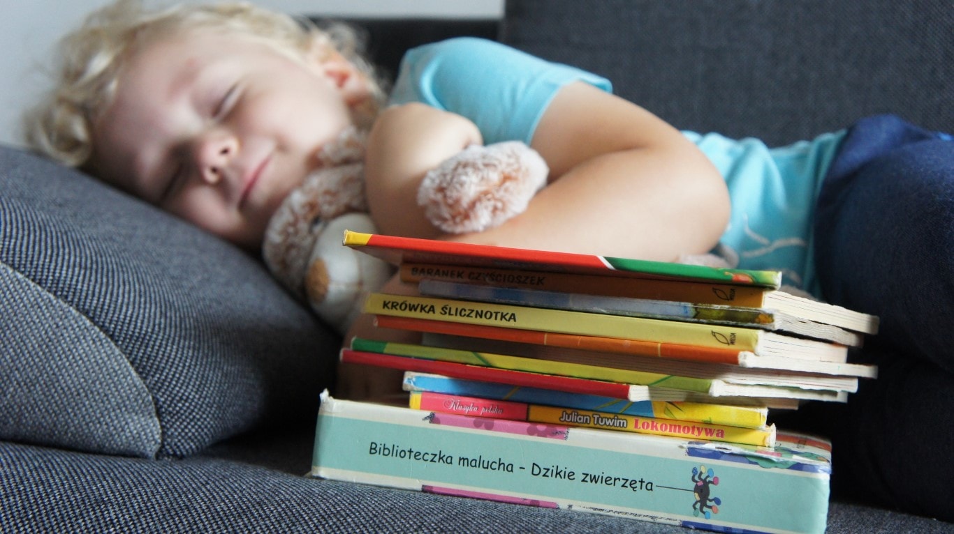Zdjęcie przedstawia dziecko śpiące na kanapie. Dziecko przytula misia. Przed maluchem nim leży stos książek.