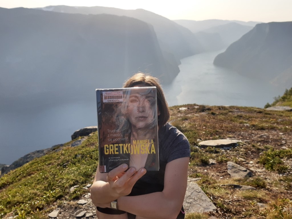 Zdjęcie przedstawia kobietę. Trzyma ona książkę, która zasłania jej twarz (sleeveface). W tle góry.