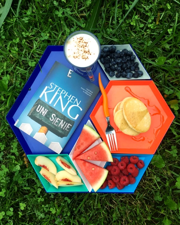 Zdjęcie przedstawia tackę z jedzeniem - borówkami, naleśnikami, malinami, arbuzem, brzoskwinią. W największej przegrodzie leży książka z kawą. W tle trawa.
