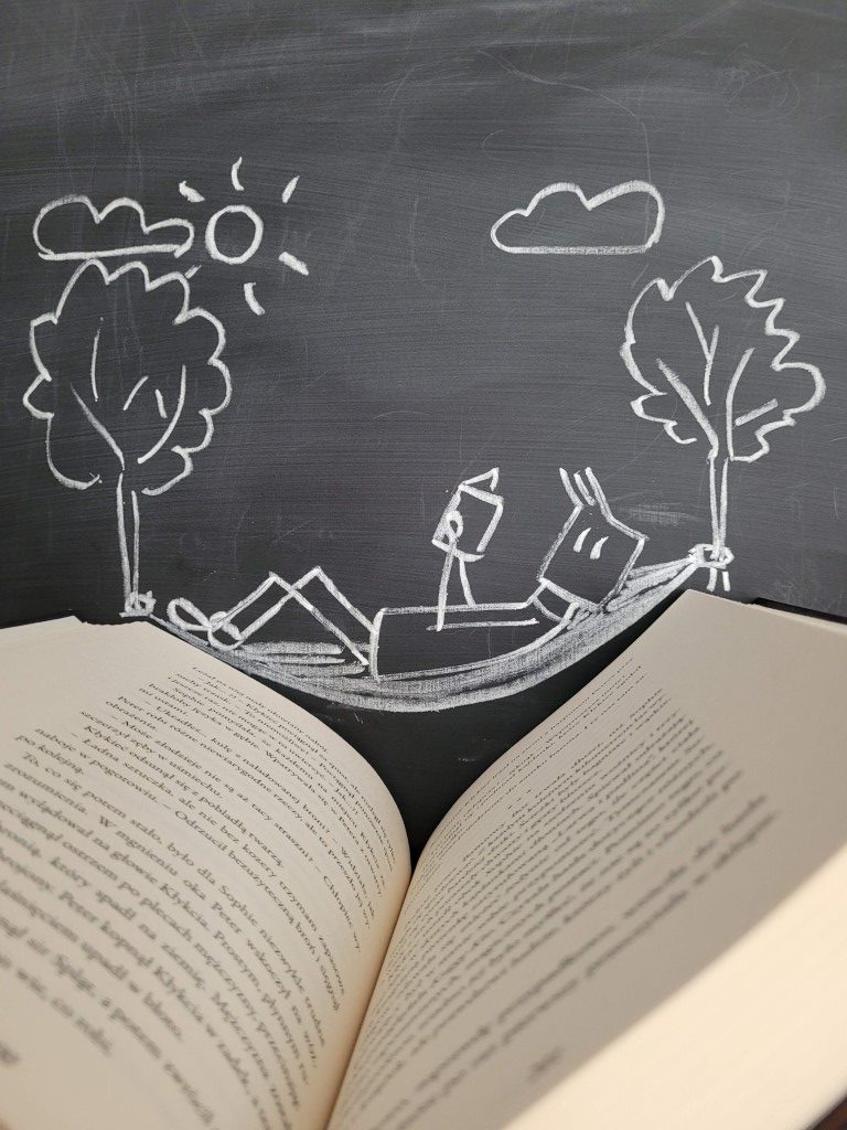 Zdjęcie przedstawia książkę. W tle czarna tablica a na niej narysowany ludzik na hamaku zawieszonym na drzewach.