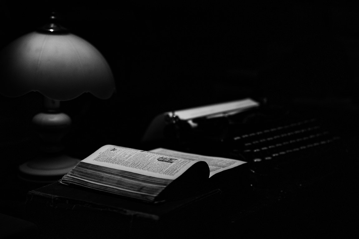 Na zdjęciu widać maszynę do pisania, książkę i lampkę. Fotografia jest czarno-biała.