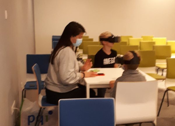 Na zdjęciu widać dwójkę dzieci korzystających z okularów VR, siedzą przy stole razem z osobą pomagającą w obsłudze.
