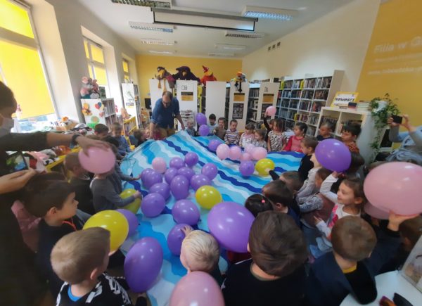 Dzieci trzymają niebieską płachtę materiału, na której umieszczono kolorowe dmuchane baloniki