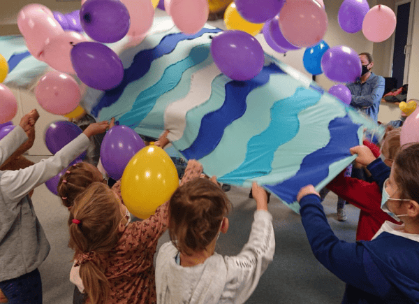 Dzieci trzymają niebieską płachtę materiału, wokół spadają kolorowe baloniki