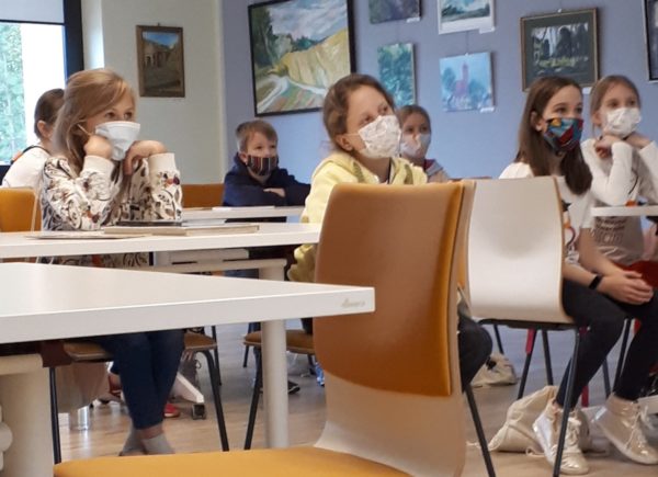 Zdjęcie przedstawia grupę dzieci siedzących na żółtych krzesłach, za białymi stołami, wszystkie dzieci mają na twarzach maseczki i widać po nich, że są mocno zasłuchane