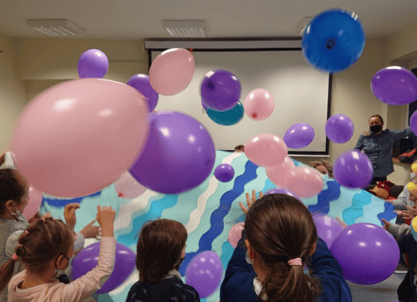 Dzieci trzymają niebieską płachtę materiału, wokół spadają kolorowe baloniki