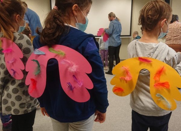 Trzy dziewczynki mają przyklejone do pleców kolorowe skrzydełka z papieru ozdobione piórkami