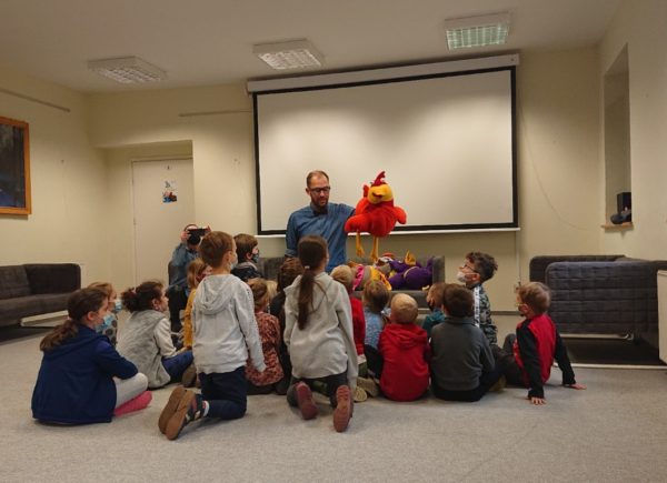 Dzieci siedzą na podłodze i słuchają pana, którzy trzyma w ręku maskotkę dużej czerwonej kury