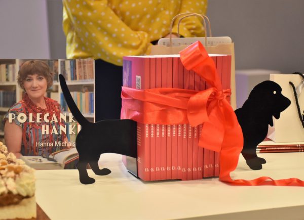 Na zdjęciu jest widoczny plik książek "Polecanki Hanki". Książki są owinięte czerwoną wstążką. Między książkami została umieszczona plastikowa figura psa rasy jamnik.