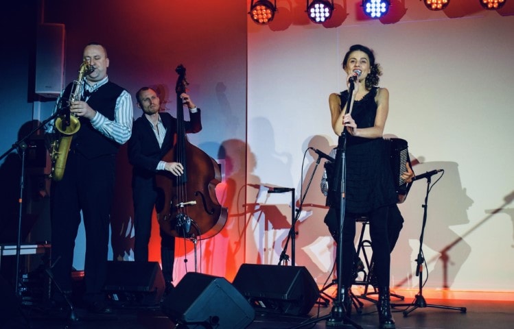 Na zdjęciu widzimy Justynę Jary i Alegancką Kapelę. Kobieta śpiewa do mikrofonu. W tle scena wraz z dwoma muzykami - trębaczem i wiolonczelistą.