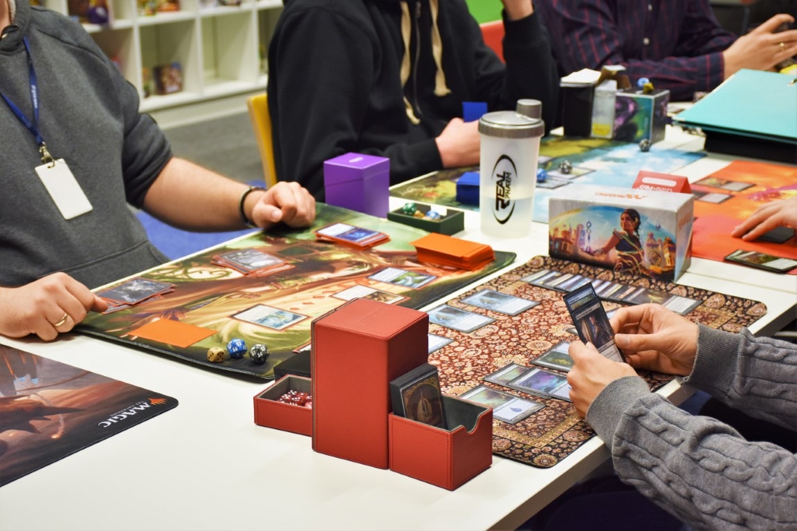 Na zdjęciu widać ręce uczestników biorących udział w turnieju Magic the Gathering w formacie Pauper. Możemy dostrzec karty i plansze gry.