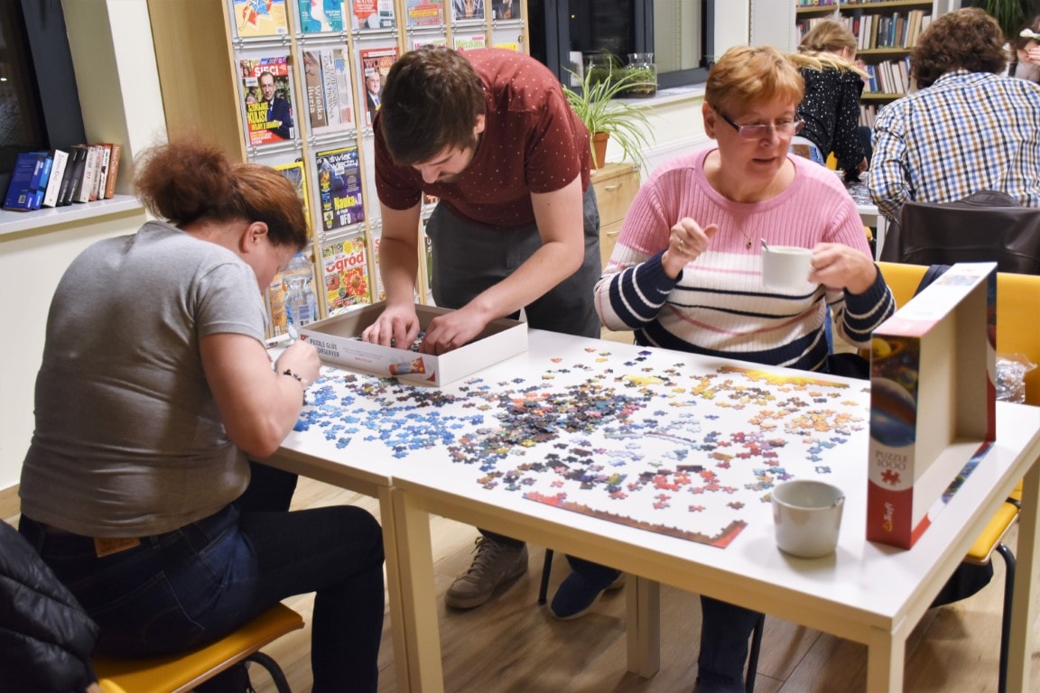 Na zdjęciu widać grupę osób: dwie kobiety i mężczyznę. Układają oni puzzle. Jedna z kobiet trzyma w ręku filiżankę.