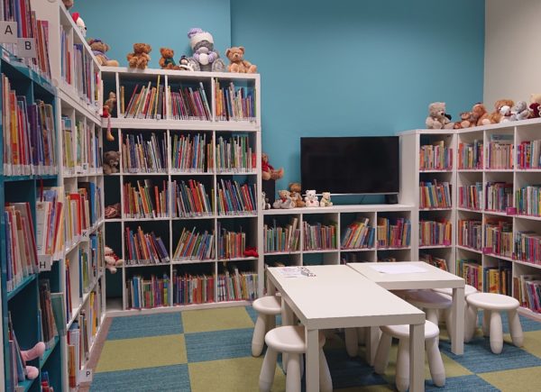Pokoik z ksiązkami dla dzieci. Na środku pomieszczenia białe stoliki. Na regale z lewej strony siedzą pluszowe misie