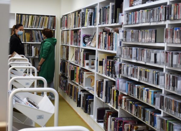 Na zdjęciu widać regały z książkami w bibliotece w Töölö. Uczestniczki projektu stoją obok półek z publikacjami.