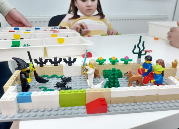 Dziewczynka układa klocki lego. Na pierwszym planie kolorowa scena z klocków