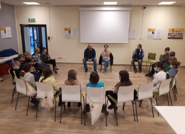 Zdjęcie przedstawia siedzących w półokręgu na krzesłach wykładowców i studentów Piaseczyńskiego Uniwersytetu Dziecięcego