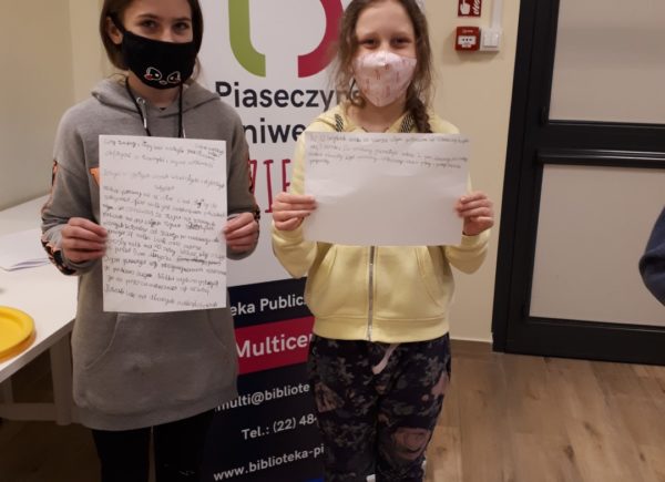 Dwie nastolatki stojące przed banerem z napisem Piaseczyński Uniwersytet Dziecięcy prezentujące swoją pracę grupową