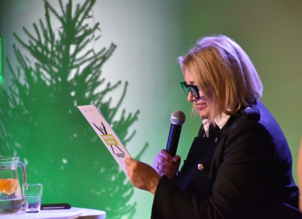 Małgorzata Ostrowska-Królikowska stoi na scenie, trzyma w ręku mikrofon i czyta treść dyktanda.