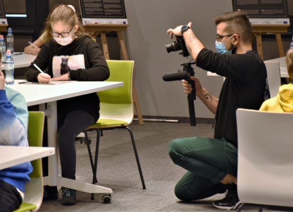 Uczestniczka konkursu siedzi przy stoliku i pisze dyktando. Po jej prawej stronie kuca fotograf i robi zdjęcie.