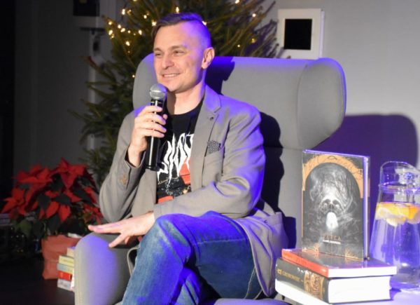 Kamil Staniszek siedzi na scenie na fotelu. Trzyma w ręku mikrofon i patrzy się w stronę publiczności. Mężczyzna uśmiecha się.