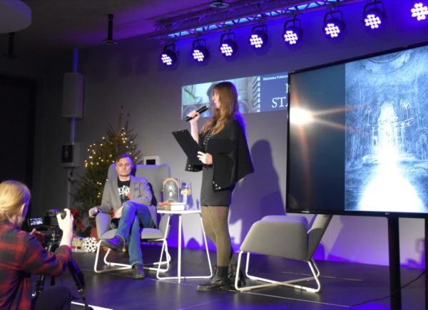 Osoba prowadząca wywiad trzyma w ręku mikrofon. Po lewej stronie widać Kamila Staniszka. Autor siedzi na fotelu i patrzy się w stronę publiczności.