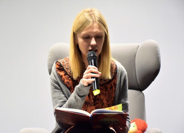Magdalena Górska siedzi na fotelu na scenie. W ręku trzyma mikrofon i czyta książkę "Aliaszka".