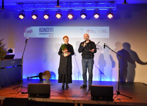 Na scenie stoją organizatorzy Koncertu Mikołajkowo-Gwiazdkowego Dobrej Woli. Mężczyzna trzyma w ręku mikrofon i przemawia do publiczności.
