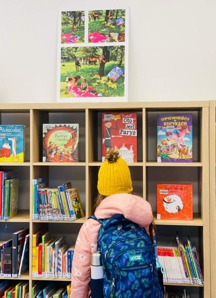 Zdjęcie przedstawia fotografię w ramach wystawy pokonkursowej "Wakacje z książką 2021". Przed tablicą z ekspozycją widać dziewczynkę, czytelniczkę punktu bibliotecznego w Kamionce.