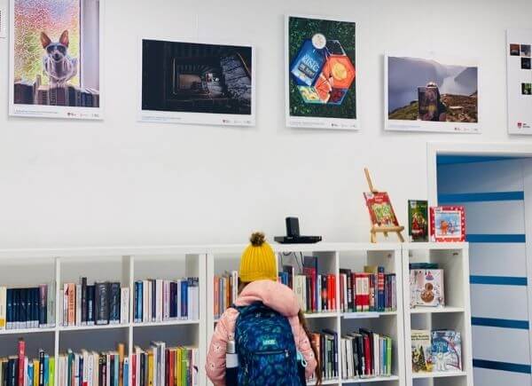 Zdjęcie przedstawia fotografie w ramach wystawy pokonkursowej "Wakacje z książką 2021". Przed tablicami z ekspozycją widać dziewczynkę, czytelniczkę punktu bibliotecznego w Kamionce.