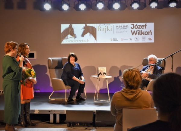 Józef Wilkoń siedzi na fotelu na scenie z mikrofonem w ręku. Obok niego na fotelu siedzi osoba przeprowadzająca wywiad. Trzyma ona w ręku mikrofon. W lewym górnym rogu stoją dwie kobiety.
