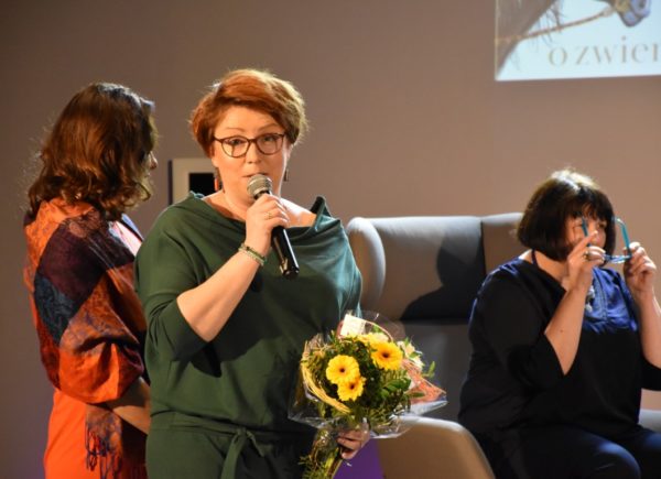 Sylwia Chojnacka - Tuzimek mówi przez mikrofon i trzyma w ręku kwiaty. W tle dwie kobiety.
