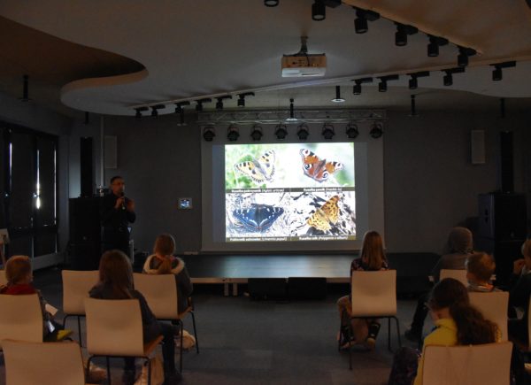 Sebastian Bielak prowadzi wykład. Widać prezentację, na której umieszczono zdjęcia motyli. Studenci siedzą i słuchają prelekcji.