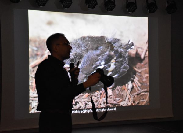 Sebastian Bielak trzyma w ręku mikrofon i aparat. W tle widać wyświetlaną prezentację.