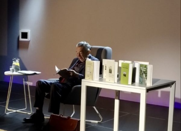 Widok na scenę, w fotelu Antoni Pieńkowski czytający fragment wiersza. Na pierwszym planie książki autora.