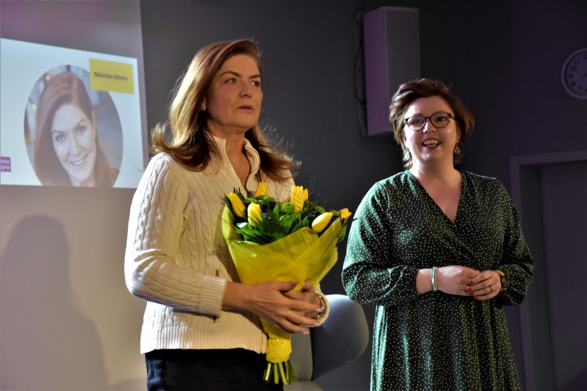 Katarzyna Dowbor stoi na scenie z bukietem żółtych tulipanów. Po jej lewej stronie stoi i uśmiecha się zastępca dyrektora Sylwia Chojnacka-Tuzimek.