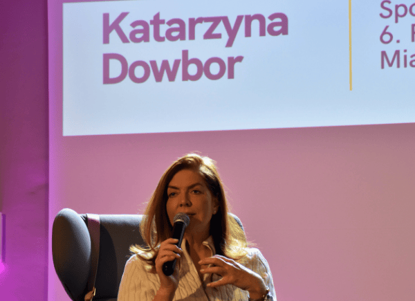 Katarzyna Dowbor siedzi na fotelu na scenie i mówi do mikrofonu. W tle różowe światła i napis promujący wydarzenie.