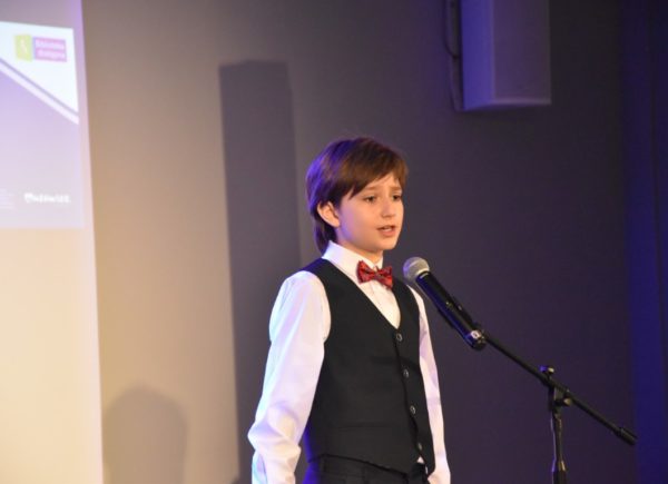 Uczestnik eliminacji stoi na scenie w garniturze. Przed nim umieszczono mikrofon. Chłopiec recytuje wiersz.