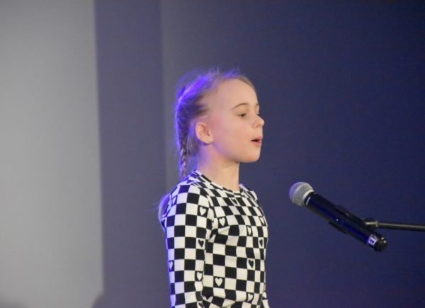 Uczestniczka eliminacji stoi na scenie w biało-czarnej sukience w kratkę. Przed nią umieszczono mikrofon. Dziewczynka recytuje wiersz.