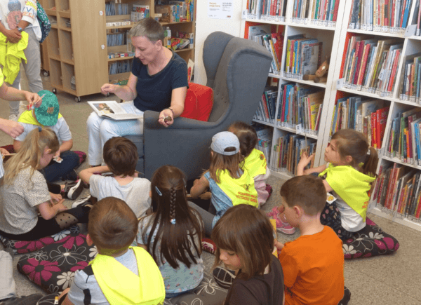 Bibliotekarka siedzi na fotelu i czyta książkę. Wokół niej siedzą dzieci