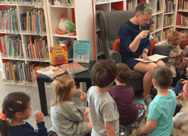 Bibliotekarka siedzi na fotelu i czyta książkę. Wokół niej siedzą dzieci