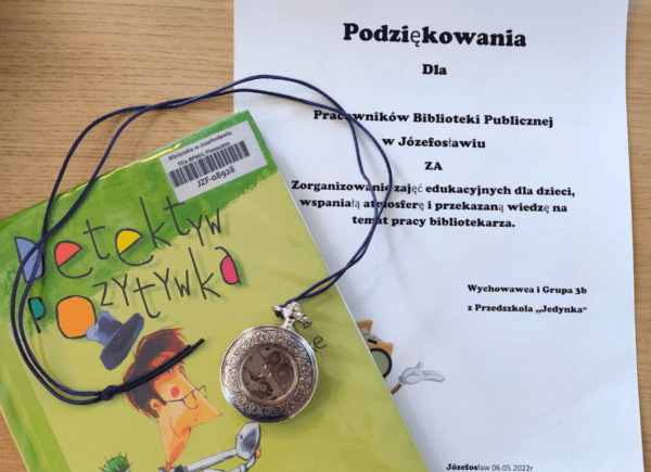 Książka „Detektyw Pozytywka”, stary kieszonkowy zegarek i kartka, na której napisane są podziękowania dla biblioteki od przedszkola